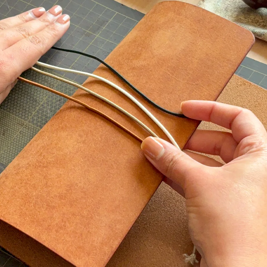 Hands laying black, cream and tan cords across a tan leather journal cover