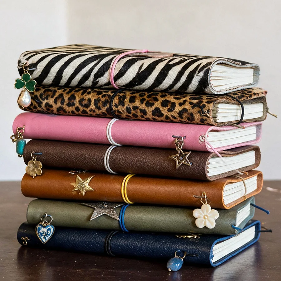 A stack of seven leather journals in zebra, leopard, pink, brown, tan, green and navy, each with charms