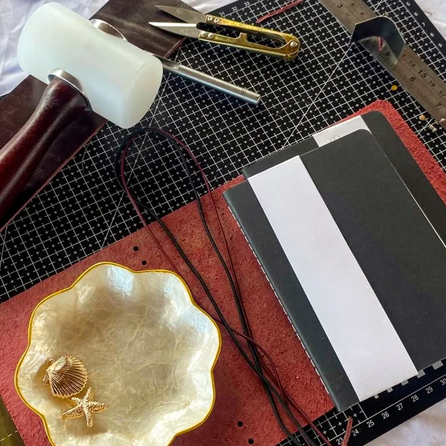 A workshop table laid out with a red leather cover, cords, journal inserts, tools and a dish of shell charms